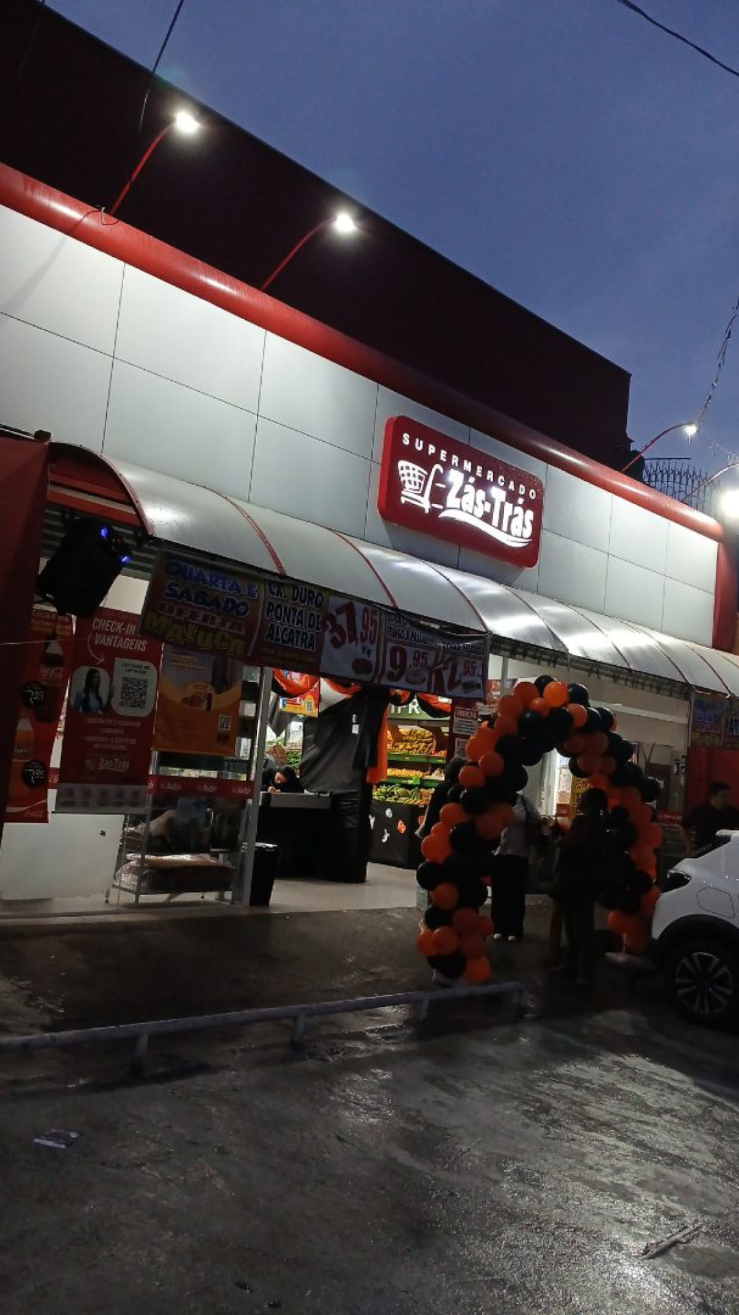 Fachada do Supermercado Zás-Trás decorada com arco de balões em dia de evento.